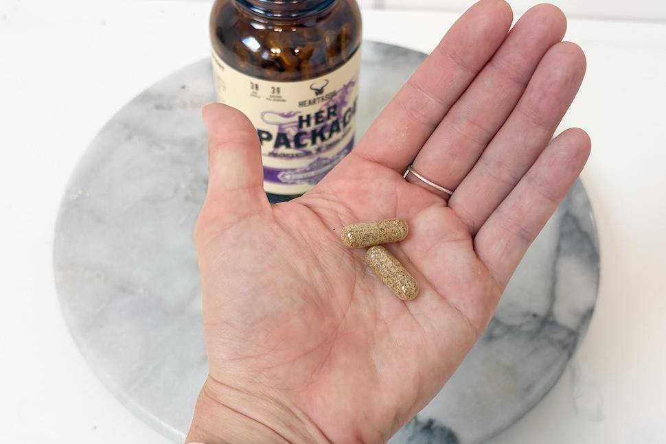An open palm holding two golden-hued pills from Heart & Soil Her Package.