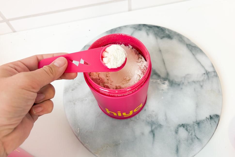 An open canister of A box of Hiya Kids Daily Growth + Protein sitting n a marble counter. The canister is open, showing the off-white powder within. Someone is using a scooper to scoop out the powder.