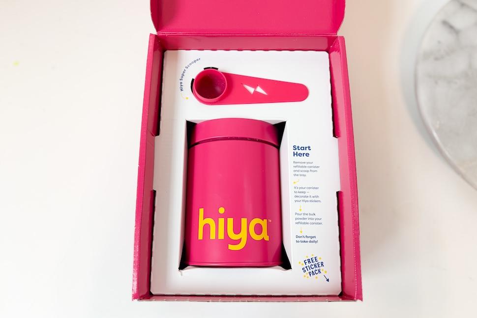 An open box of A box of Hiya Kids Daily Growth + Protein. It shows the canister of protein and scooper, both magenta in color.