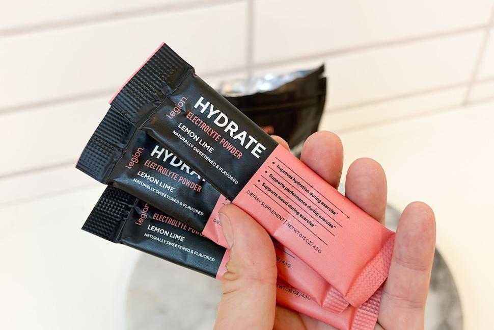 A hand holding three packets of Legion Hydrate electrolyte powder (lemon lime flavor)