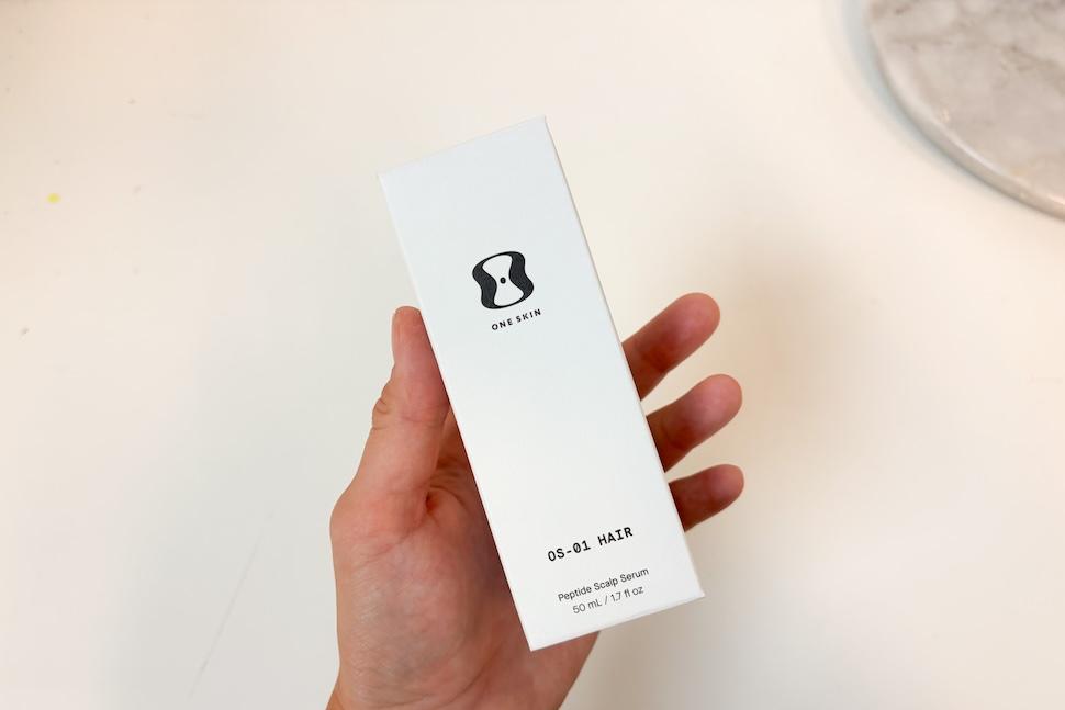 A hand holding a box of OneSkin OS-01 Hair Scalp Serum
