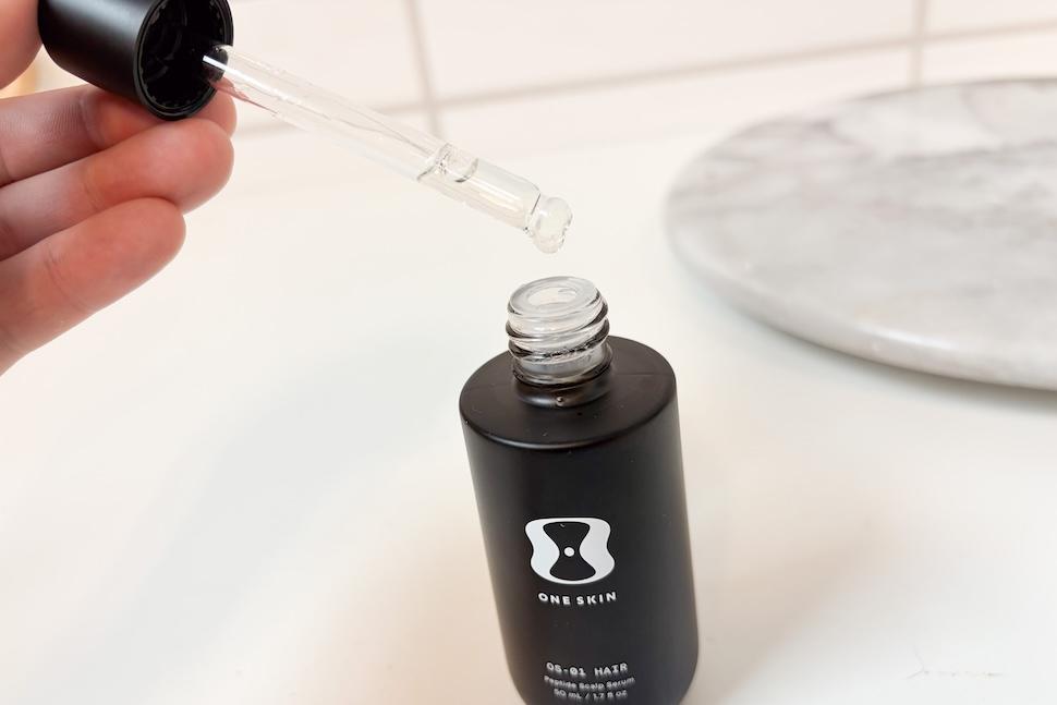 A hand extracting OneSkin OS-01 Hair Scalp Serum from its bottle with an eye dropper