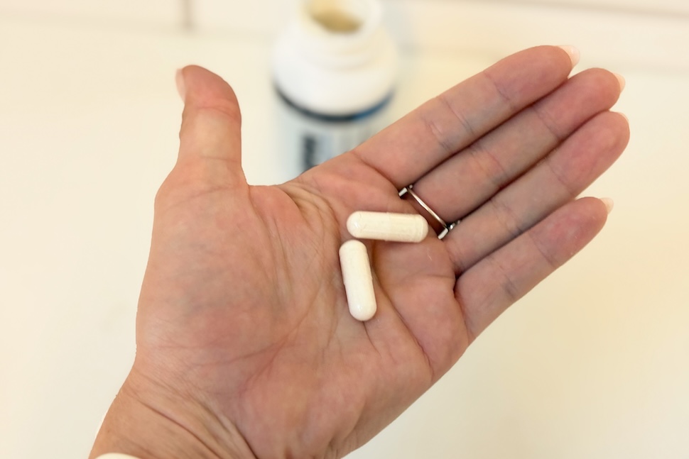 An open palm holding two Transparent Labs NMN capsules. In the background, you can see the open bottle on the counter below.