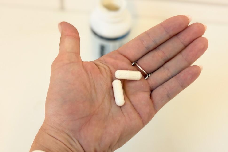 An open palm holding two Transparent Labs NMN capsules. In the background, you can see the open bottle on the counter below.