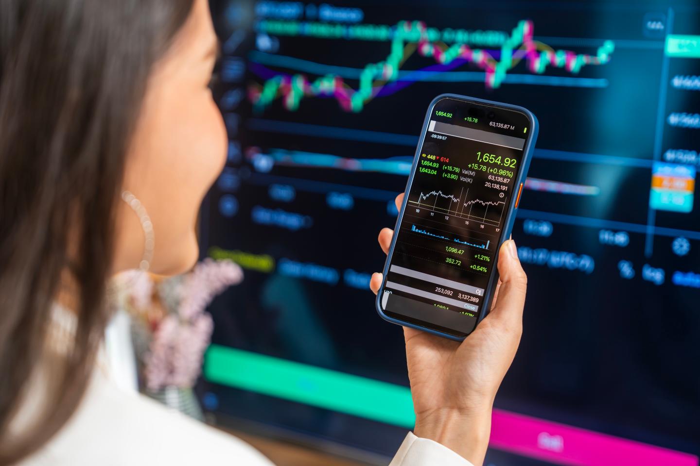 A woman observes stock trends on her smartphone while a computer screen displays financial graphs.