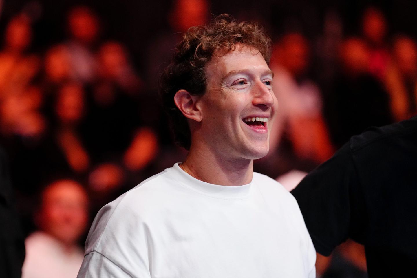 Mark Zuckerberg, wearing a white shirt, smiles. He is standing in front of a crowd.