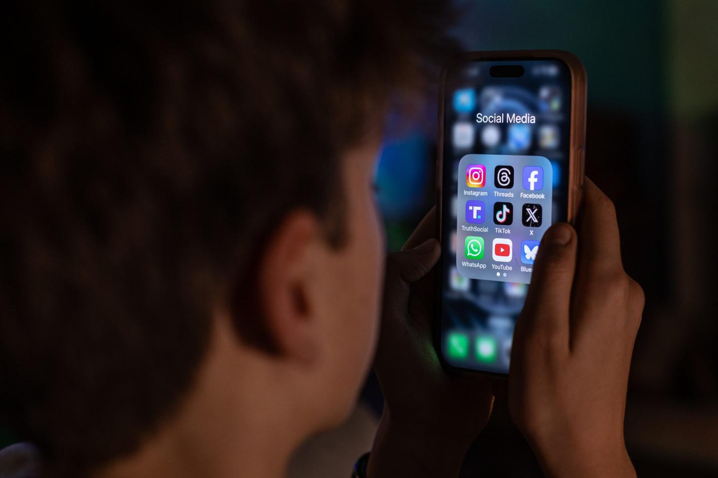 A 13-year-old teenage boy looks at an iPhone screen displaying various social media apps.
