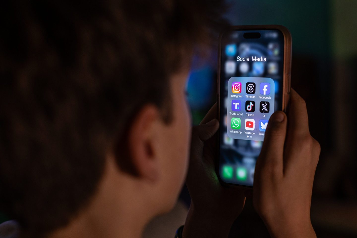 A 13-year-old teenage boy looks at an iPhone screen displaying various social media apps.