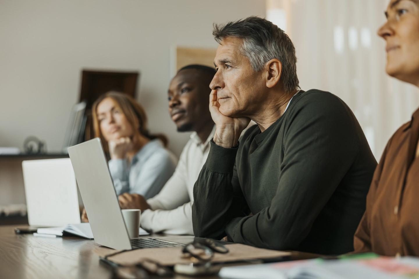 An older man sits at a table with a laptop in front of him with his chin resting on his hand.