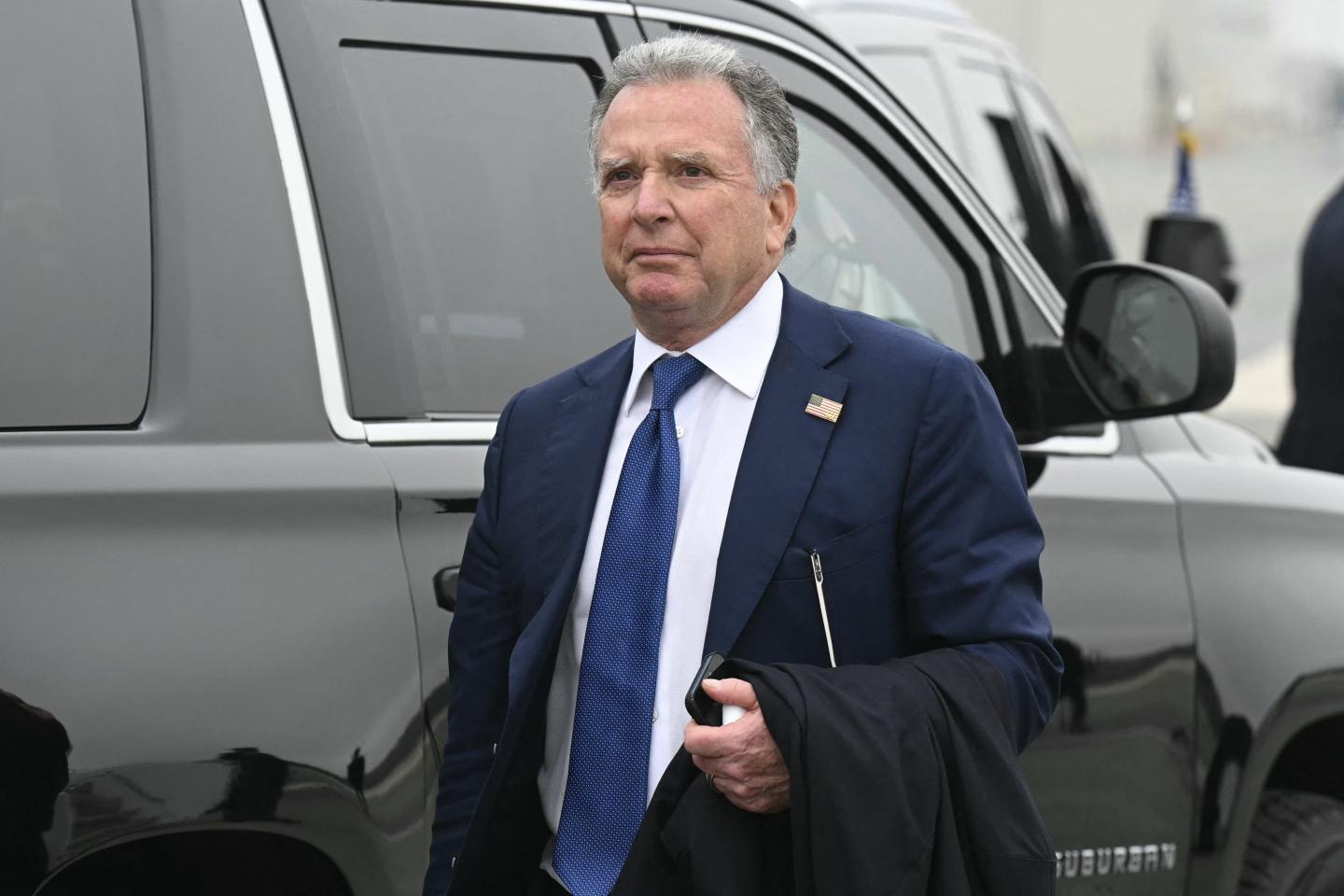 Steve Witkoff walks in front of a car dressed in suit and tie with his phone in his hand and his jacket draped over his arm.