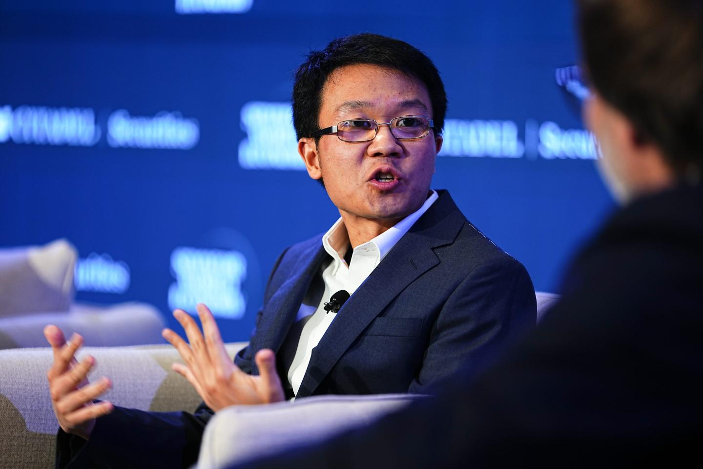 Scott Wu, in front of a blue background, sits in a gray chair and speaks to a person out of frame.