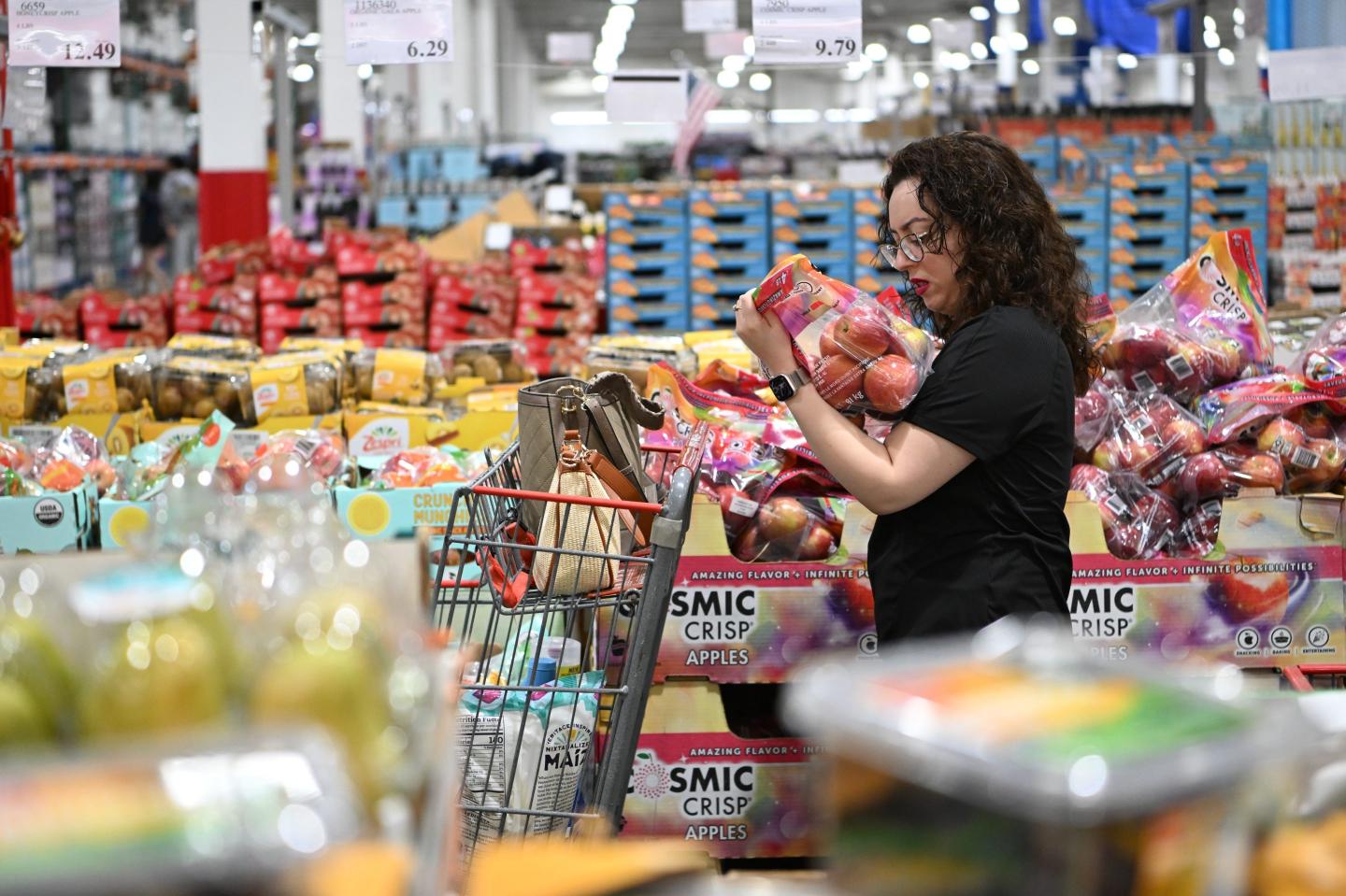 A woman, standing in a grocery store, looks at a bag of apples.