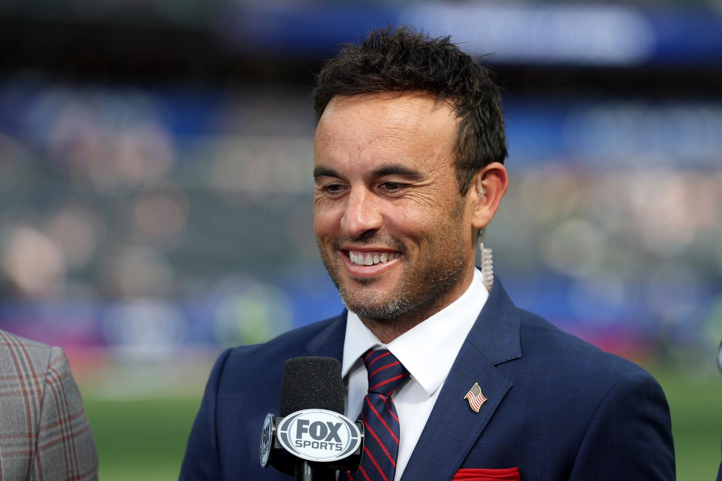 Landon Donovan, wearing a suit and holding a microphone, smiles.