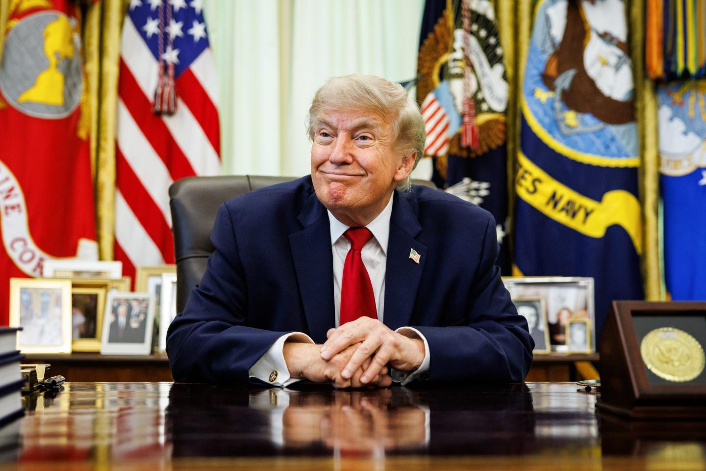 Donald Trump sits at his desk in the Oval Office, smiling and with his hands folded in front of him.