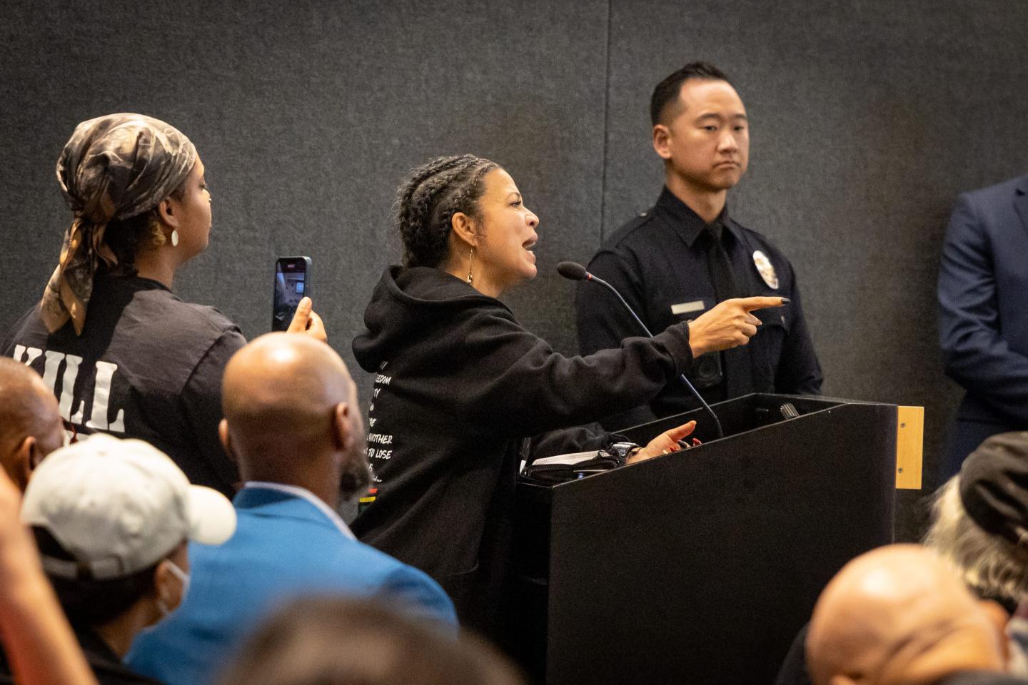 A woman at a police commission meeting stands behind a podium speaking animatedly.