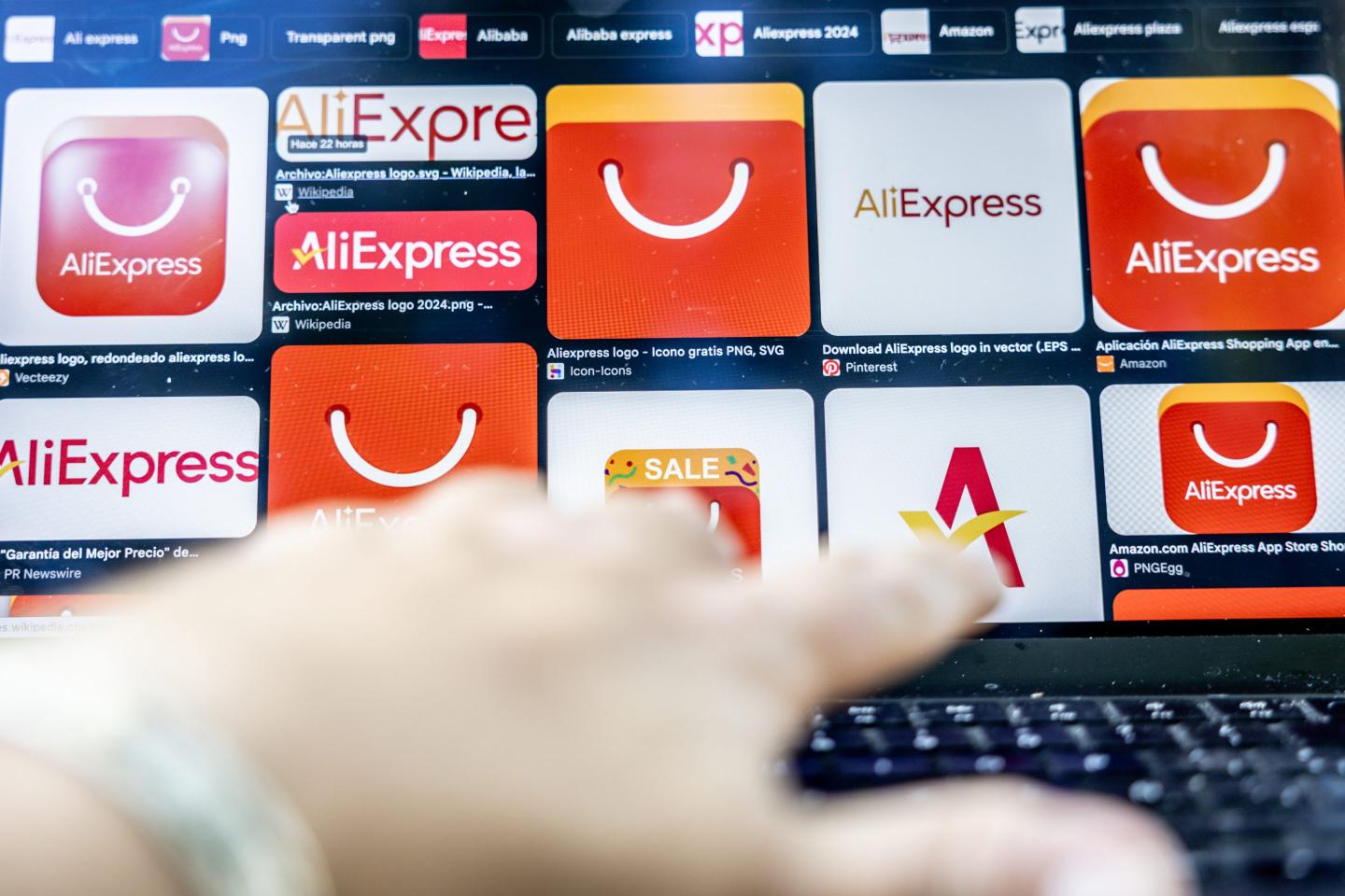 A hand types on a computer with a screen displaying the AliExpress logo from Google Images.