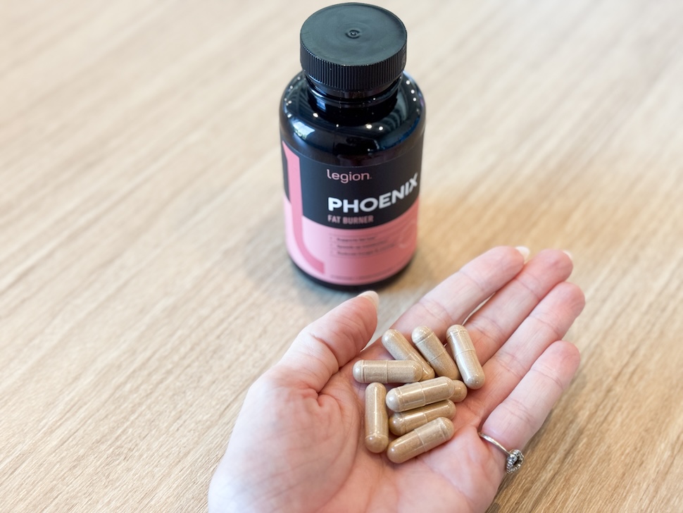 A bottle of Legion Phoenix Fat Burner sits on a wooden countertop. A hand is open next to it, showing a handful of the golden-hued capsules.