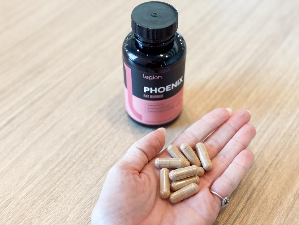 A bottle of Legion Phoenix Fat Burner sits on a wooden countertop. A hand is open next to it, showing a handful of the golden-hued capsules.