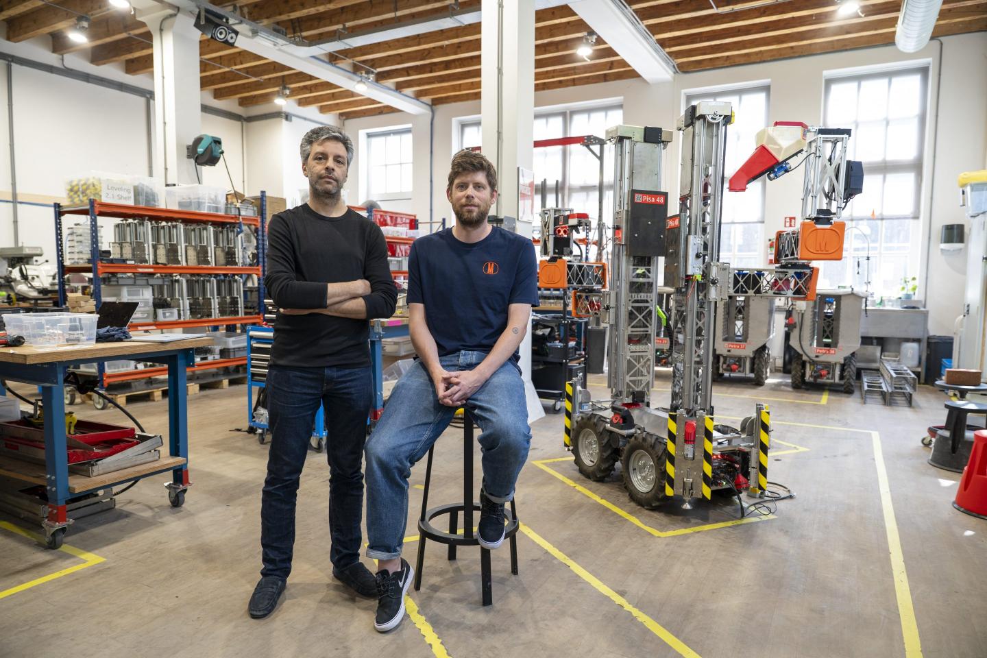 Salar al Khafaji stands while Sebastiaan Visser sits on a stool in front of a robotics warehouse