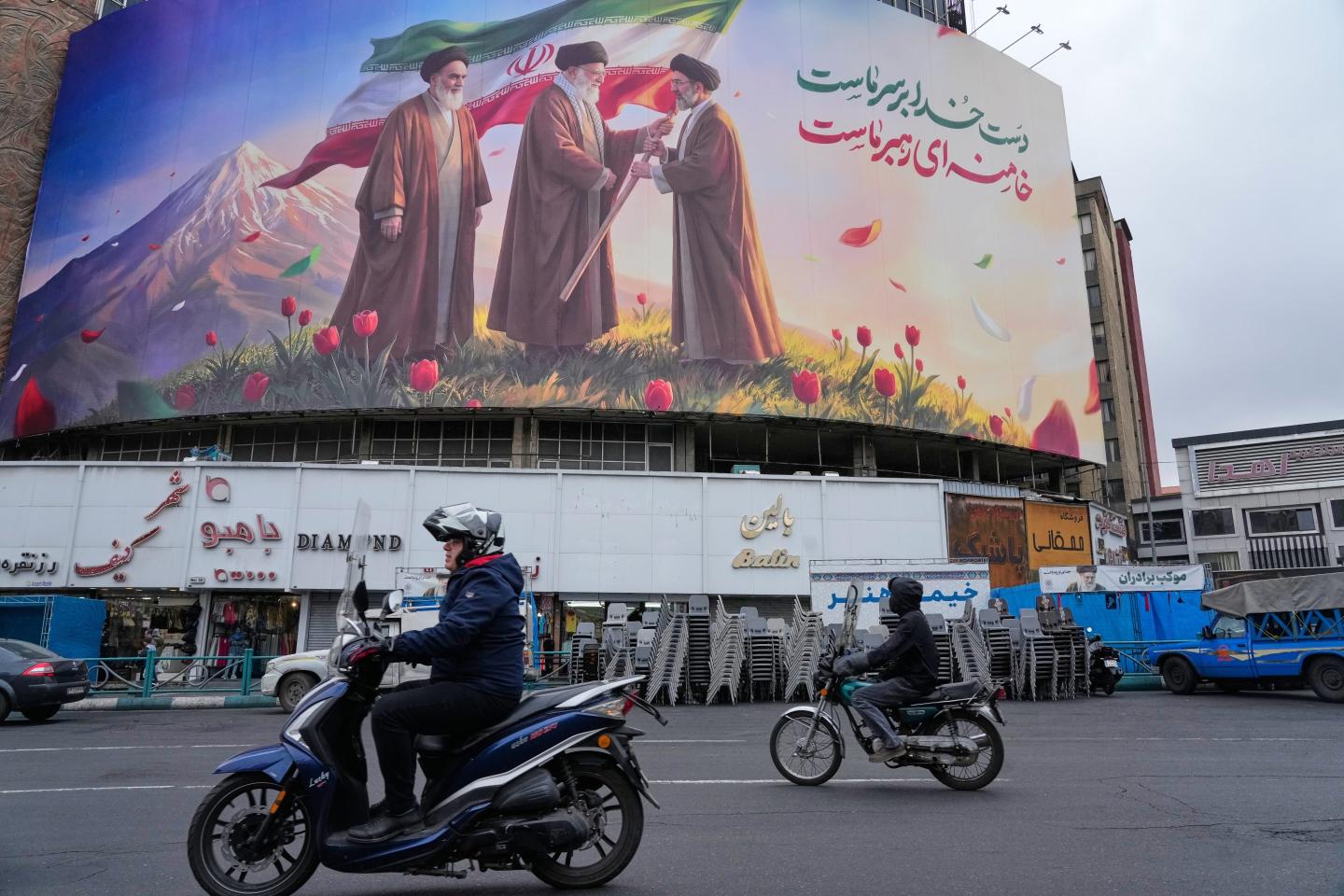 Motorbikes drive past a billboard depicting Iran's late Supreme Leader Ayatollah Ali Khamenei, center, handing the country's flag to his son and successor, Ayatollah Mojtaba Khamenei, right, as the late revolutionary founder Ayatollah Ruhollah Khomeini stands at left, in a square in downtown Tehran, Iran, March 10, 2026.