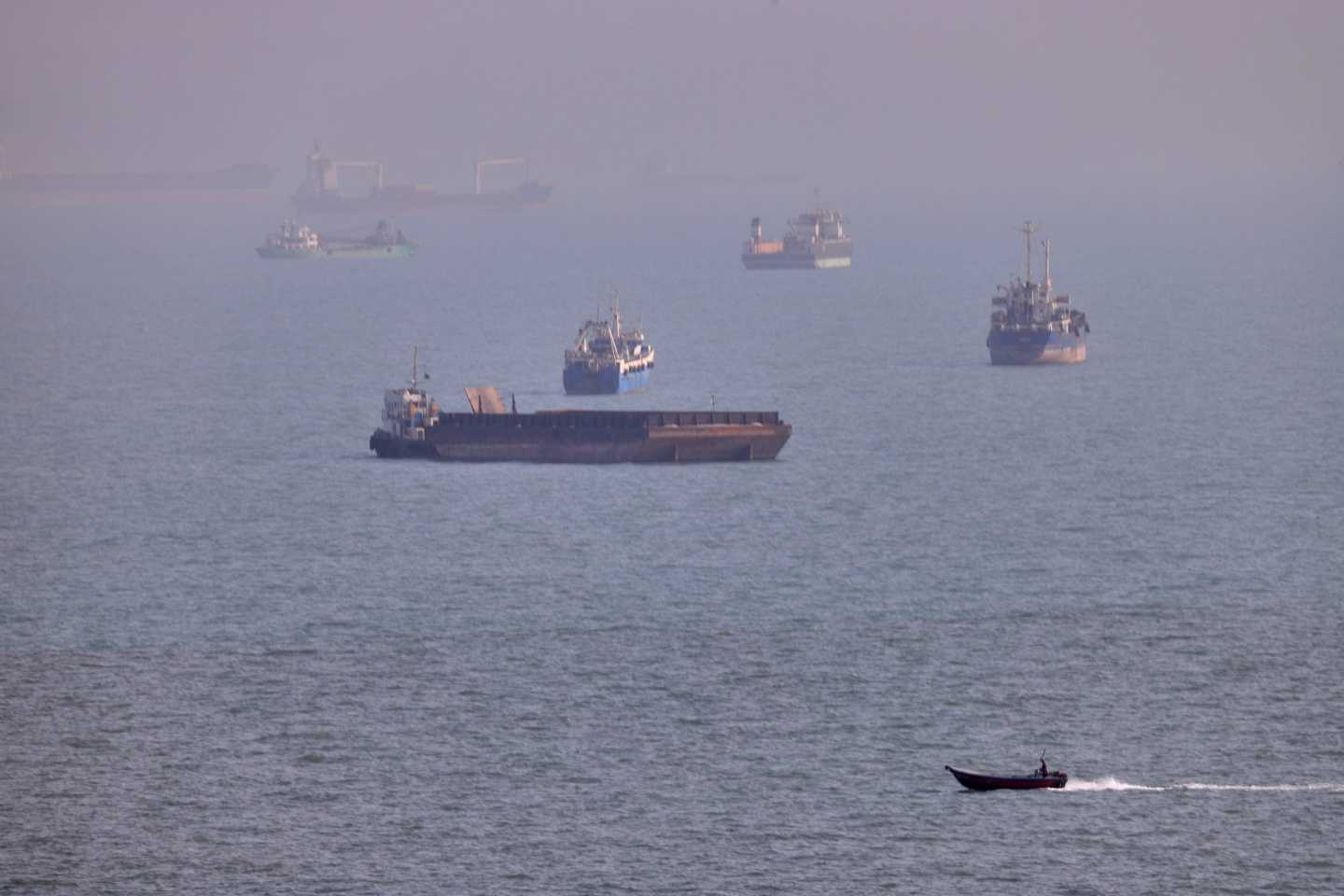 A small boat sails past cargo ships and other commercial vessels anchored in the Strait of Hormuz off Bandar Abbas, Iran.