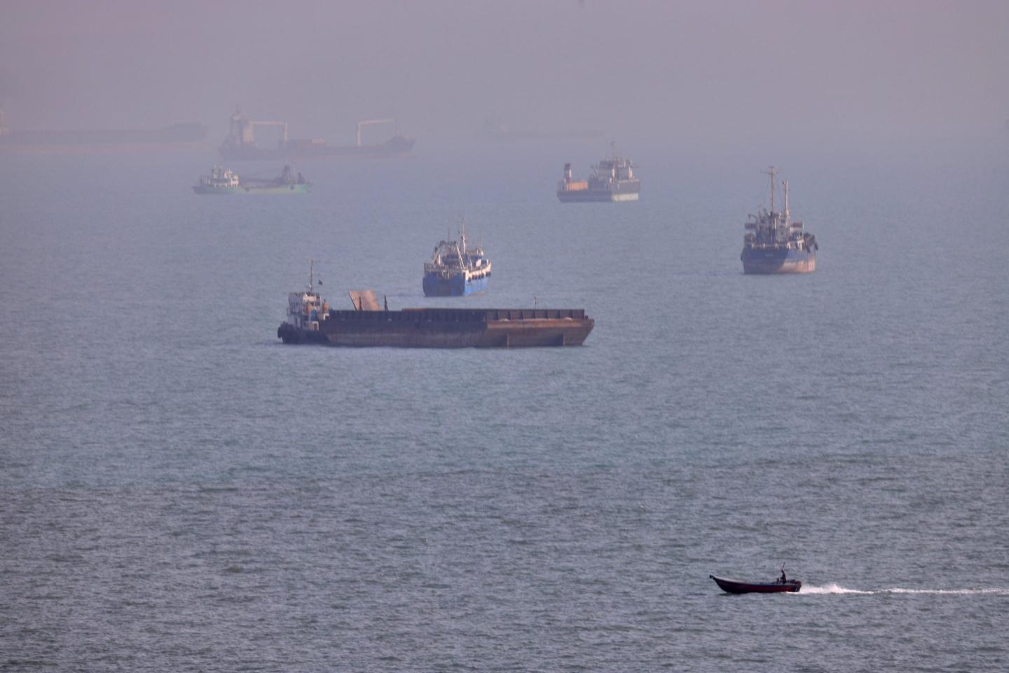 A small boat sails past cargo ships and other commercial vessels anchored in the Strait of Hormuz off Bandar Abbas, Iran.