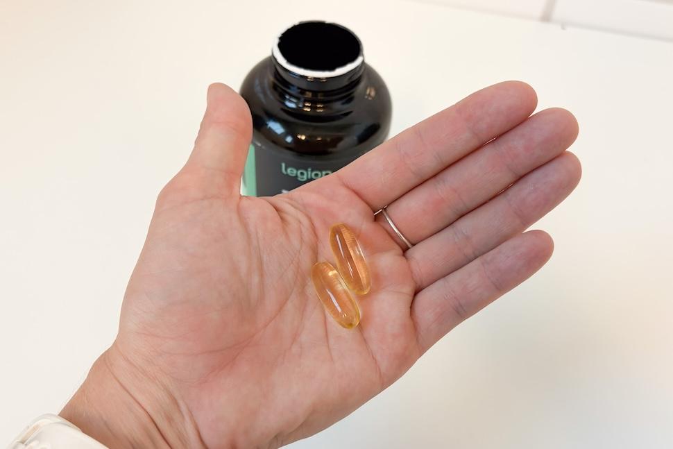 Closeup of an open palm holding two Legion Triton capsules. The capsules are a golden hue, and you can see the open bottle on the countertop below.