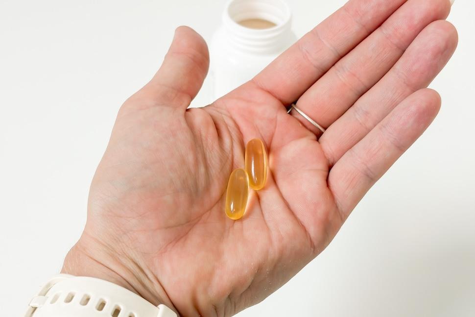 An open palm holding two Performance Lab Omega-3 capsules, which are golden in color. Below, you can see the open pill bottle on a white countertop.