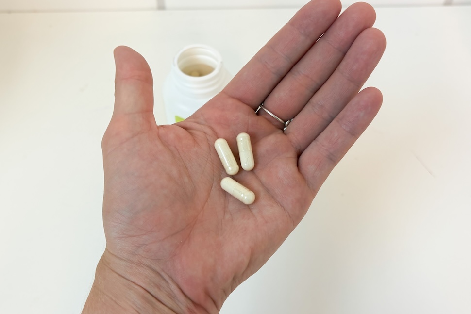 Closeup of a hand holding three Biotin Wellah capsules, which are off-white in color. You can see the open bottle on a white countertop below.