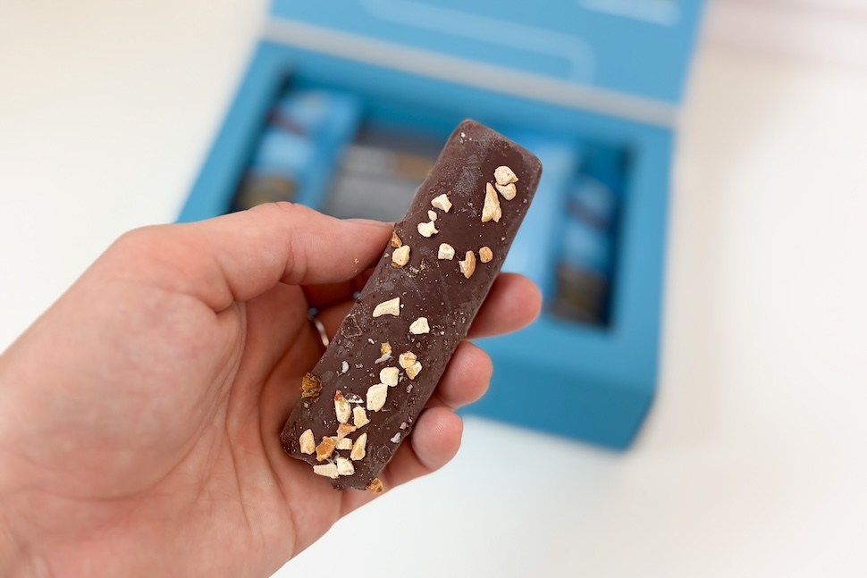 Closeup of a hand holding an unwrapped Wonderfeel ChocoCreatin bar. You can see the blue box of bars below on a white countertop.