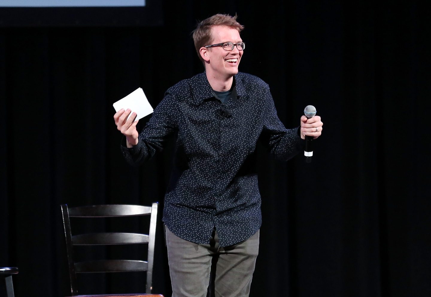 Hank Green, wearing a black shirt, stands onstage holding a card and a mic.