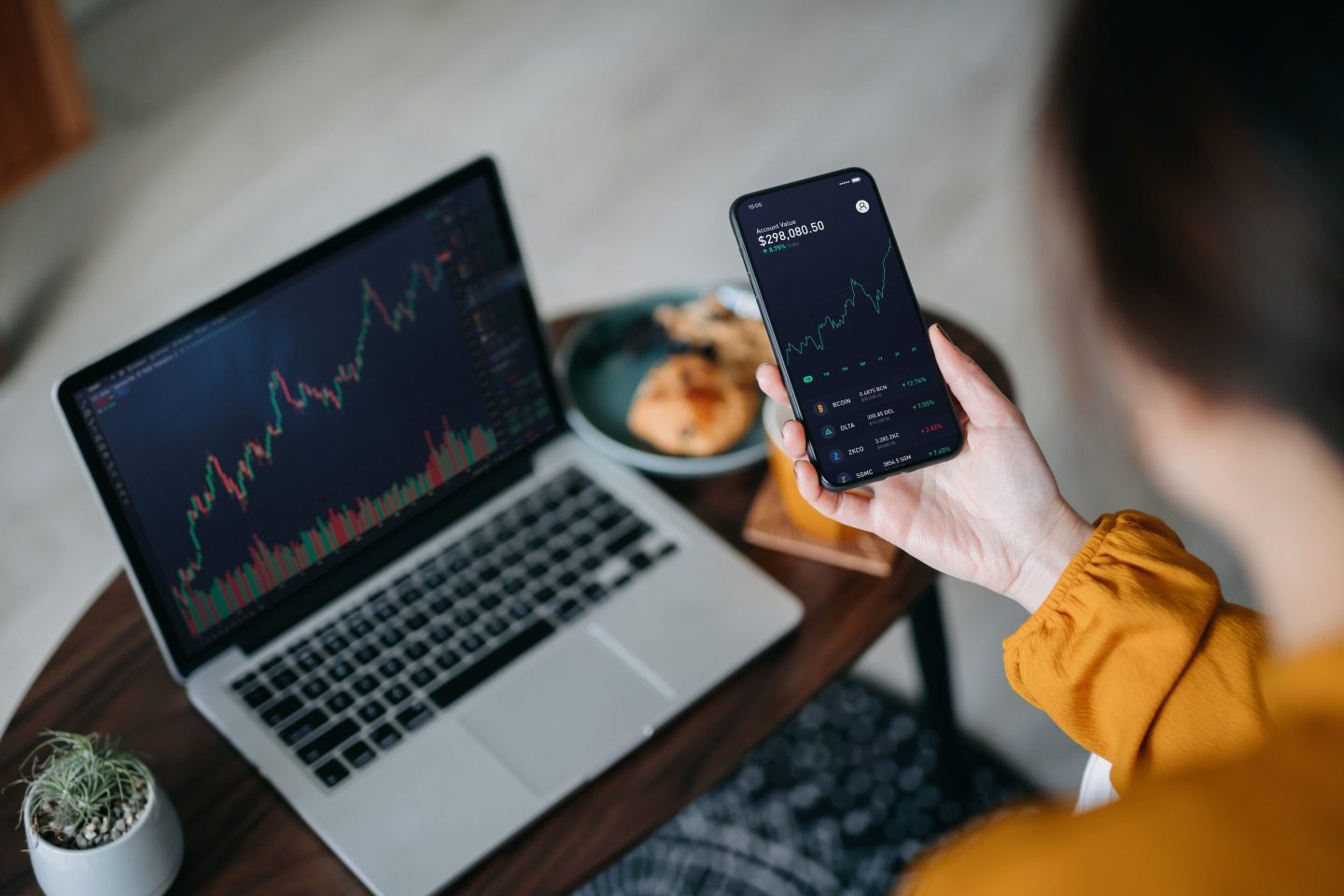 Woman holds phone displaying crypto price chart with a laptop in front of her also showing crypto price chart.
