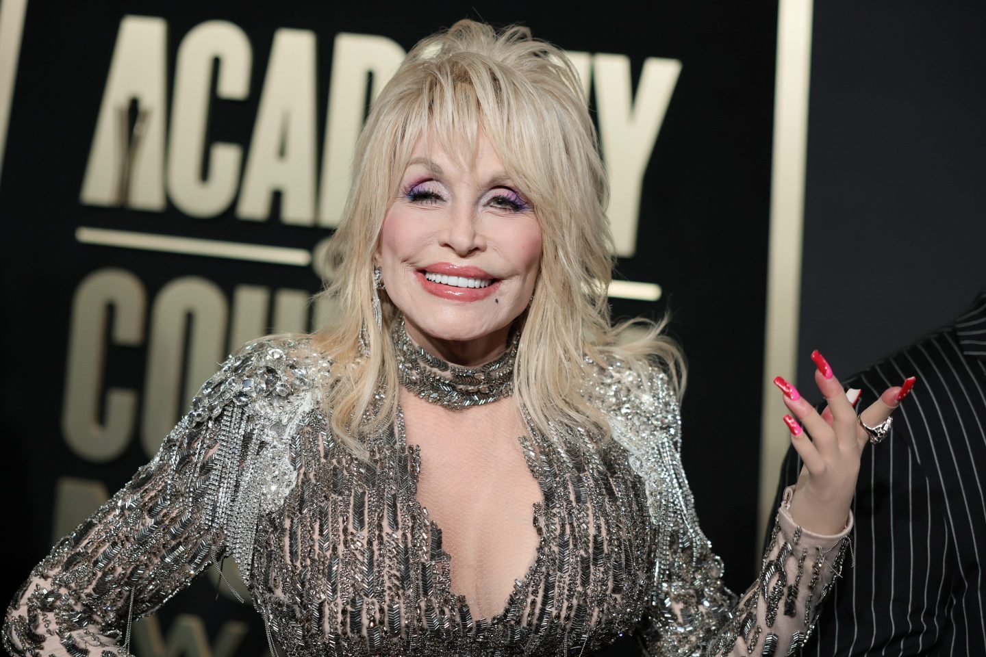Dolly Parton smiles at the camera with her hand up and fingers spread apart.