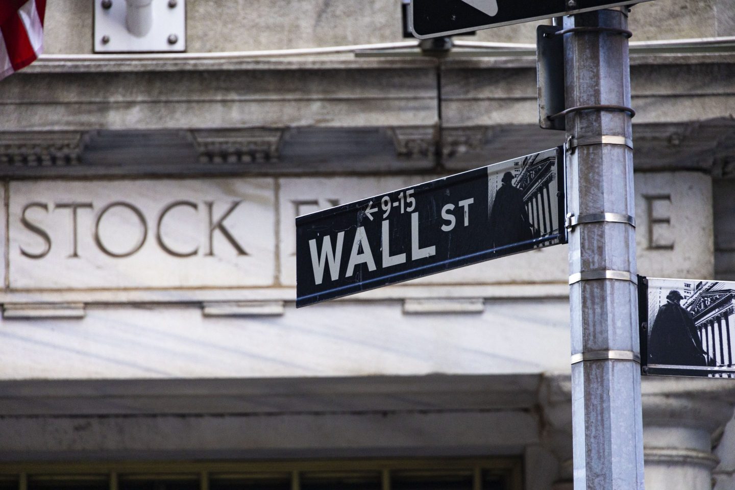 Wall Street sign inscription as seen on the road and buildings of Wall St with the entrance of New York Stock Exchange in the background.