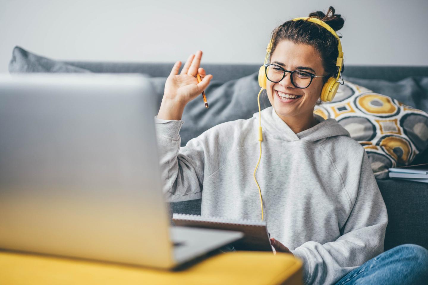 A cheerful woman in yellow headphones waves at her laptop during a video call.