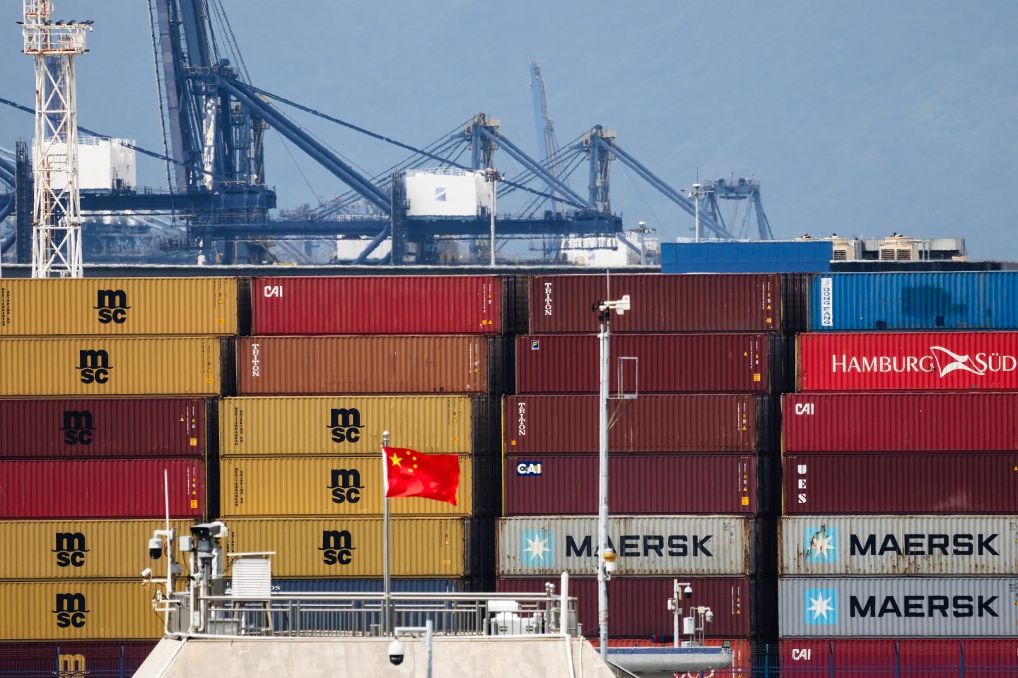 The Chinese national flag is seen in front of stacked shipping containers bearing MSC (Mediterranean Shipping Company), Maersk, and Hamburg Süd branding at Yantian Port.