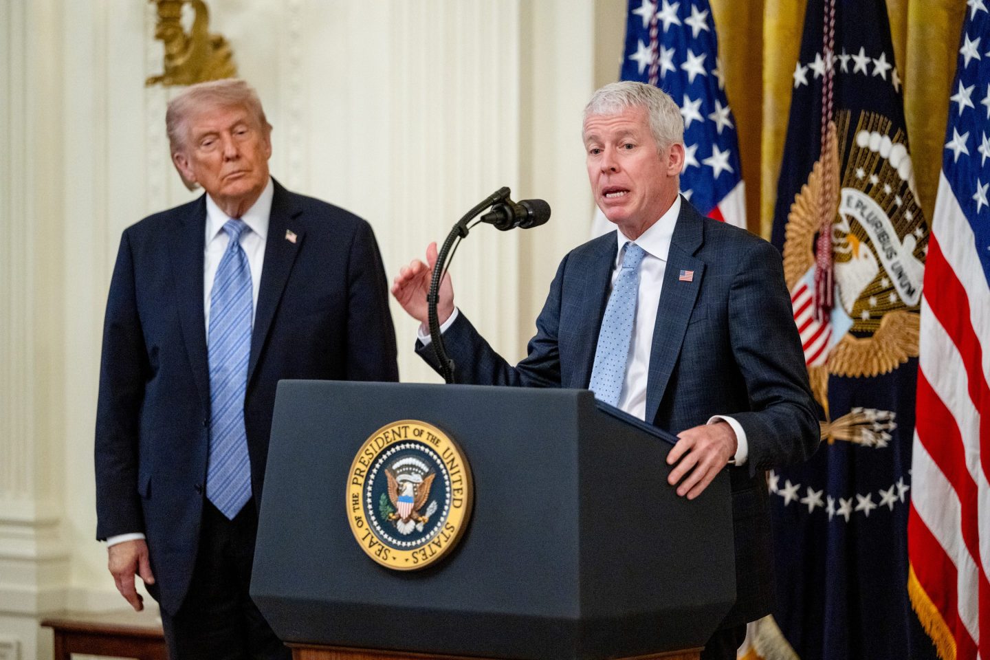 Chris Wright speaks behind a podium with Donald Trump onlooking.