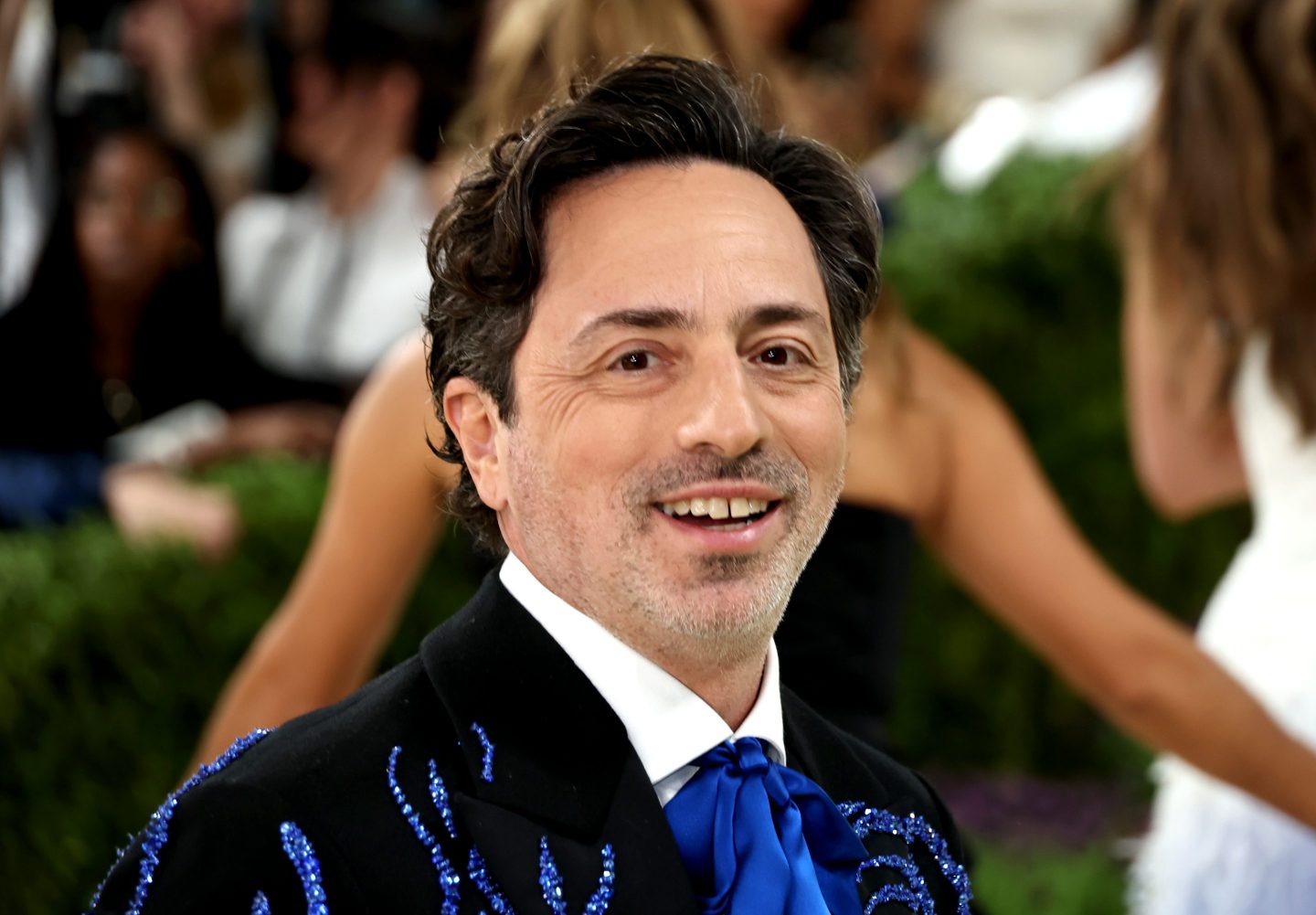 Sergey Brin, wearing a blue suit, looks at the camera and smiles.