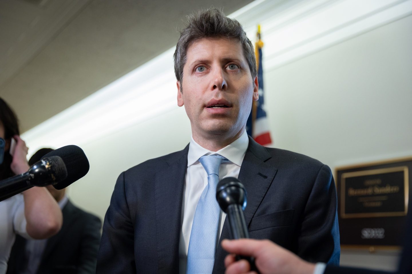 Sam Altman stands in front of microphones in a suit