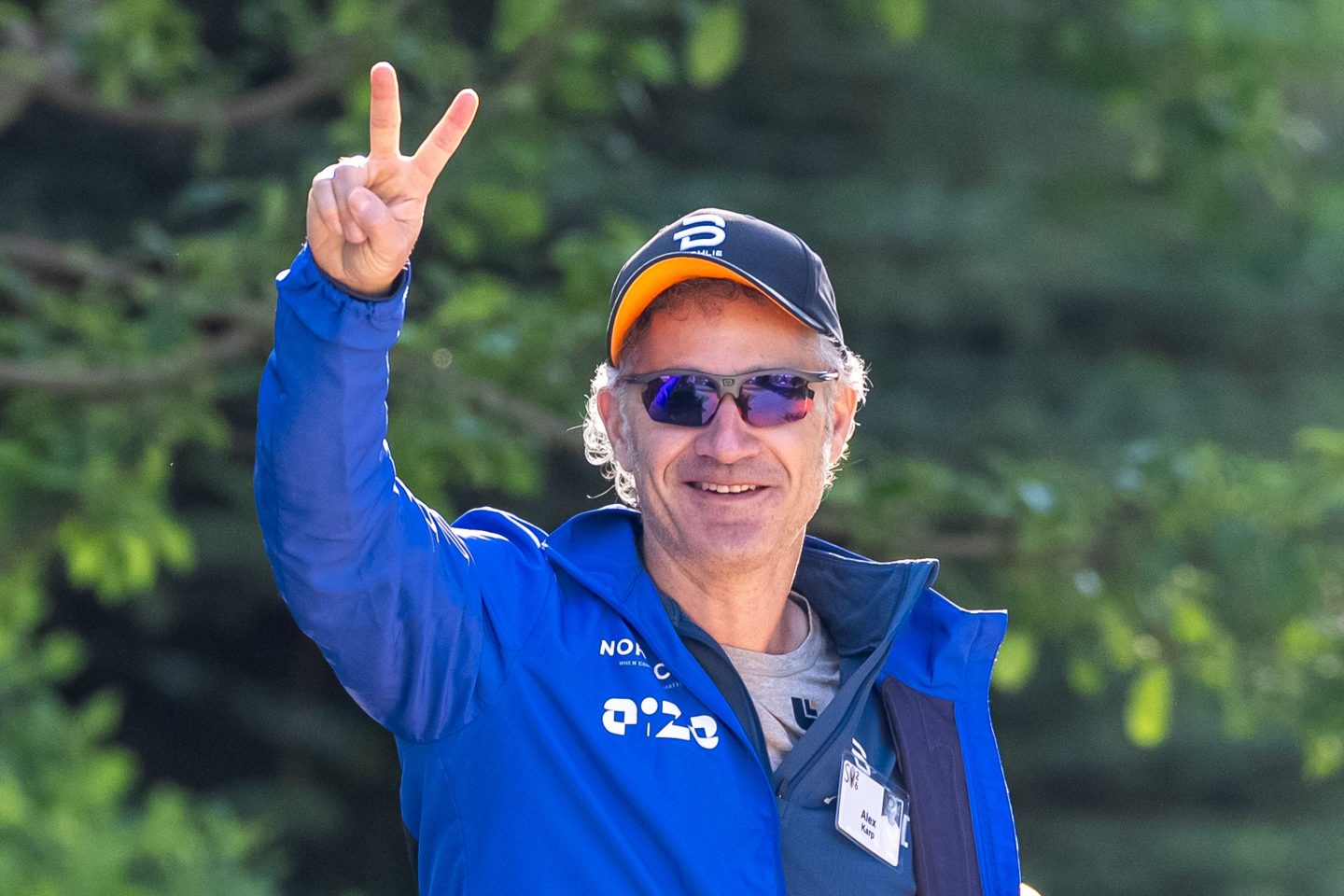 Alex Karp, wearing a hat, gives a peace sign