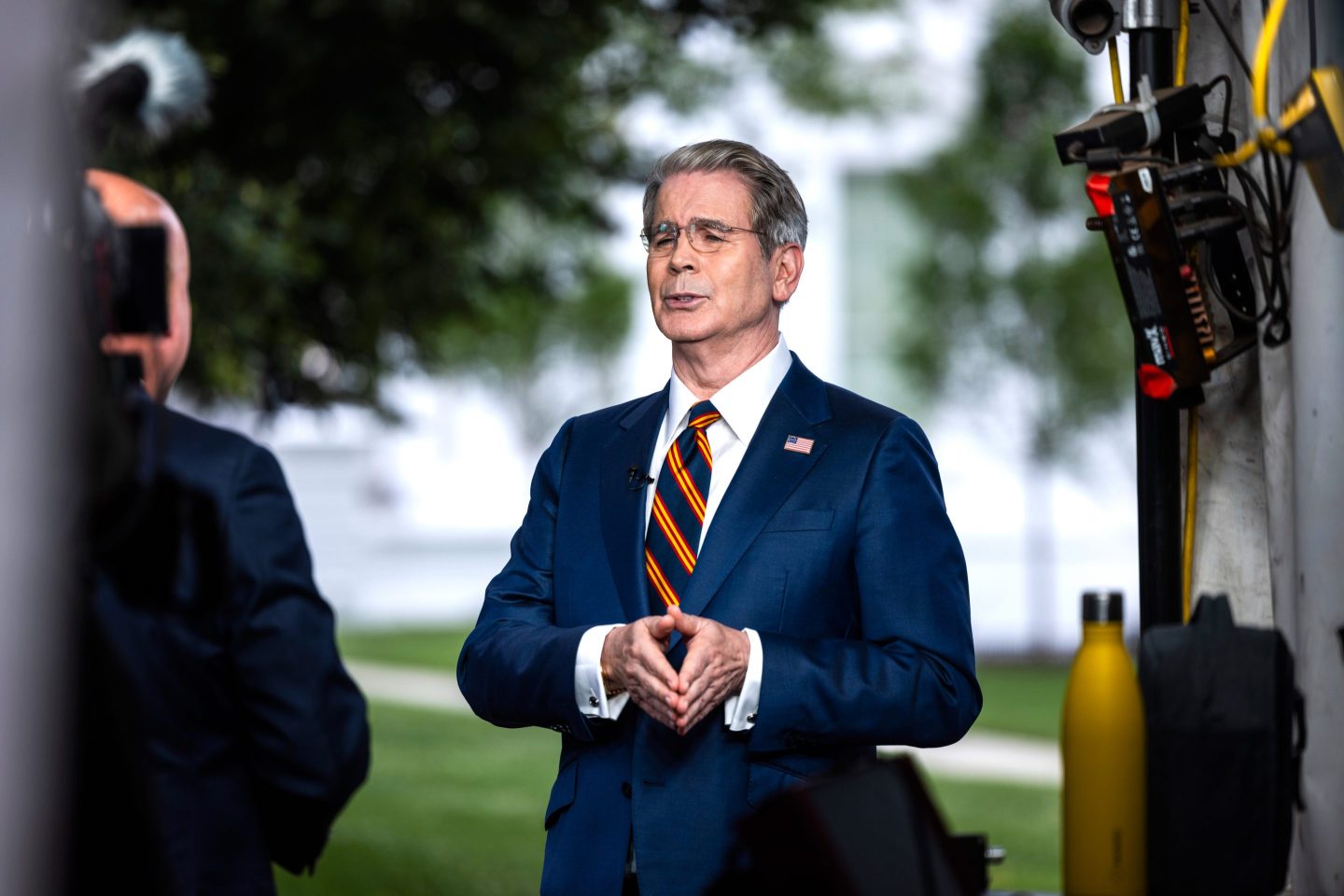 Scott Bessent stands outside wearing a suit, his hands pressed together as he speaks.