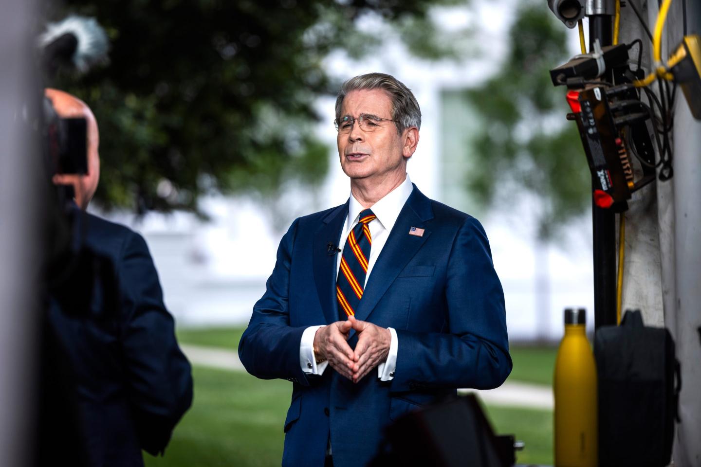 Scott Bessent stands outside wearing a suit, his hands pressed together as he speaks.
