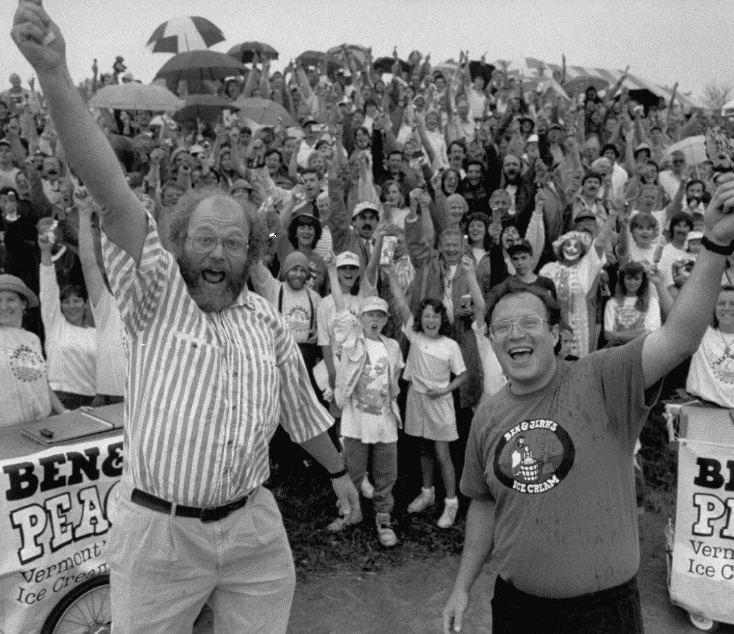 Founders Ben Cohen and Jerry Greenfield raise their arms and cheer in front of a large crowd at an outdoor Ben & Jerry's event