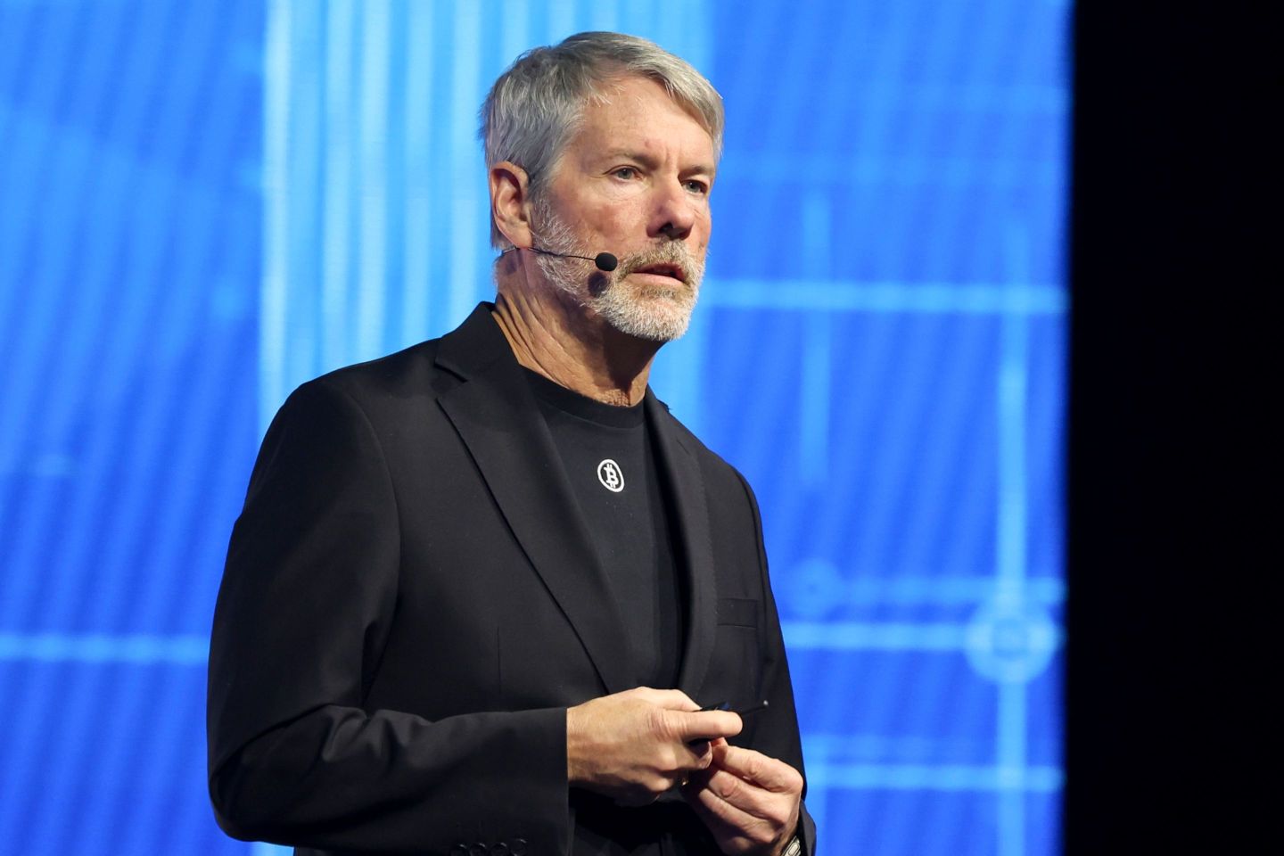 Michael Saylor with a cheek microphone, wearing a black suit, and wearing a Bitcoin pin.
