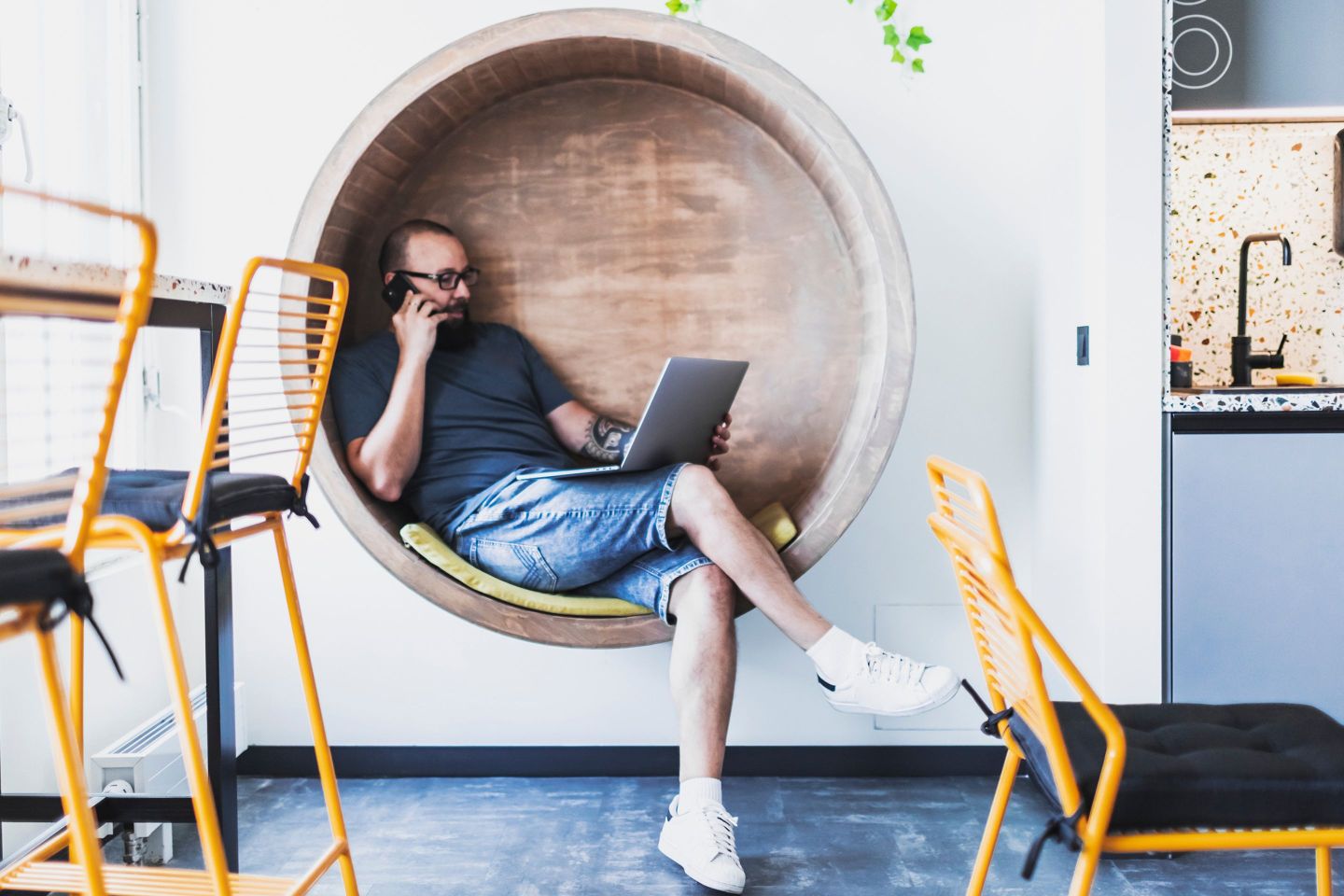 Man, wearing jeans shorts, works on a laptop and speaks on the phone in an office kitchen space.