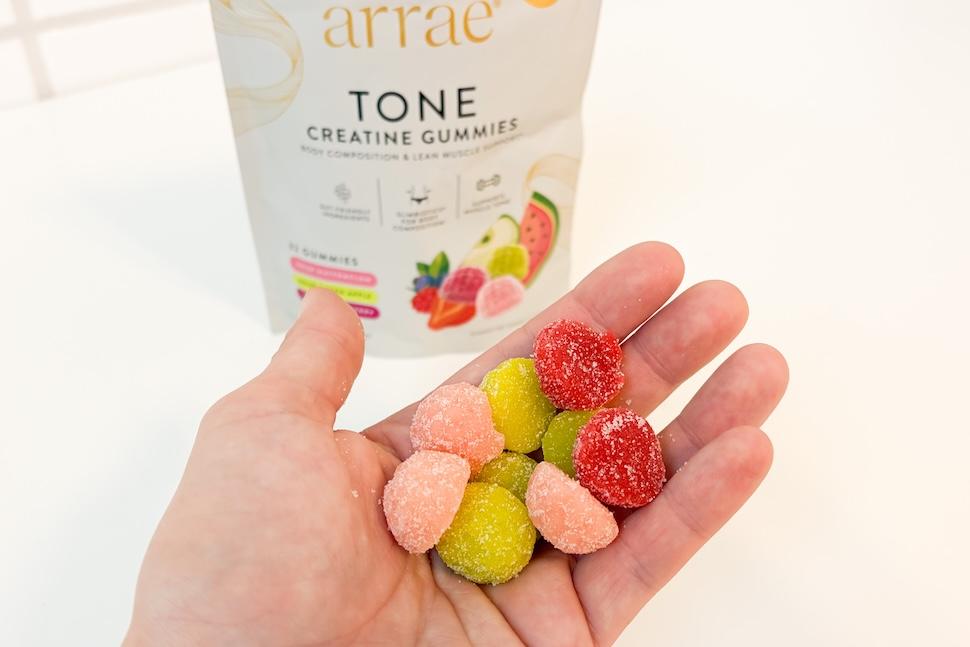 Closeup of a hand holding a multicolored variety of Tone Creatine Gummies. You can see the bag on the white countertop below.
