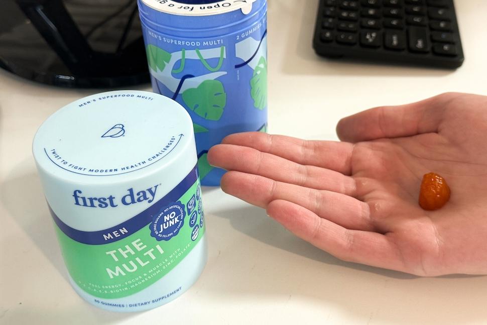 An open palm holding a First Day Men's the Multi gummy. The green and white-colored canister sits on a desk next to the hand.