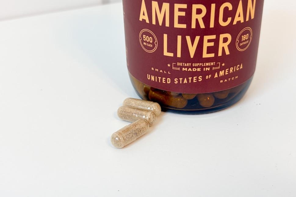 A bottle of Heart & Soil Pure American Liver sitting on a white countertop. Three brownish capsules sit next to the bottle.