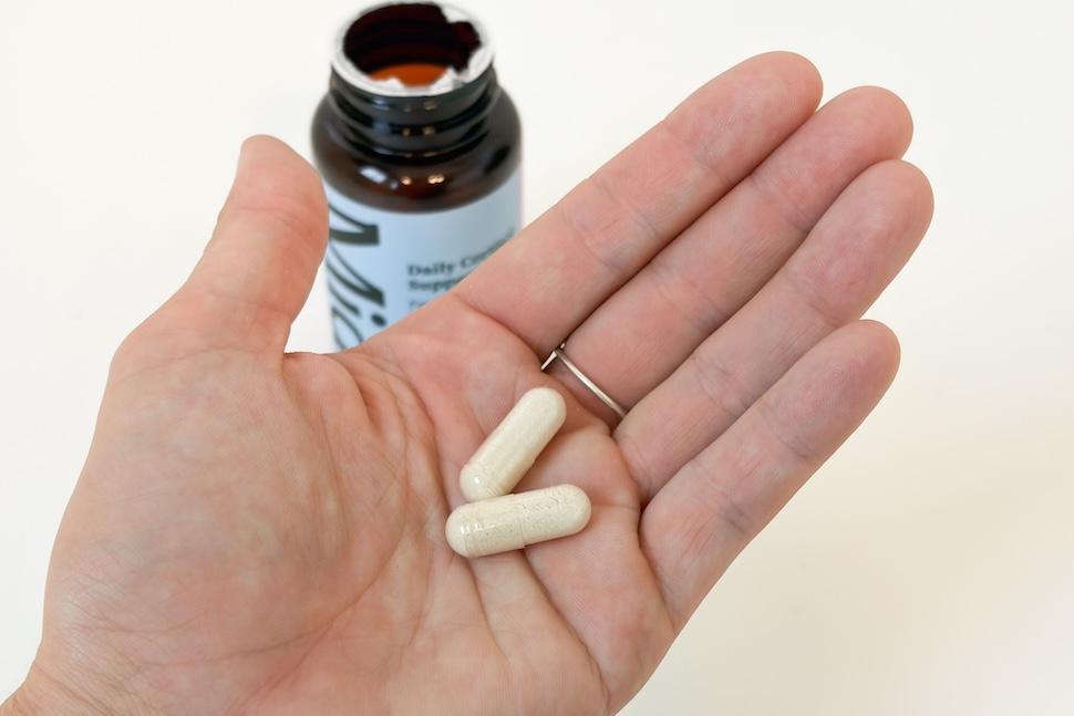 An open palm holding two off-white Midi Daily Cortisol Support capsules. Below, an open pill bottle sits on a white countertop.