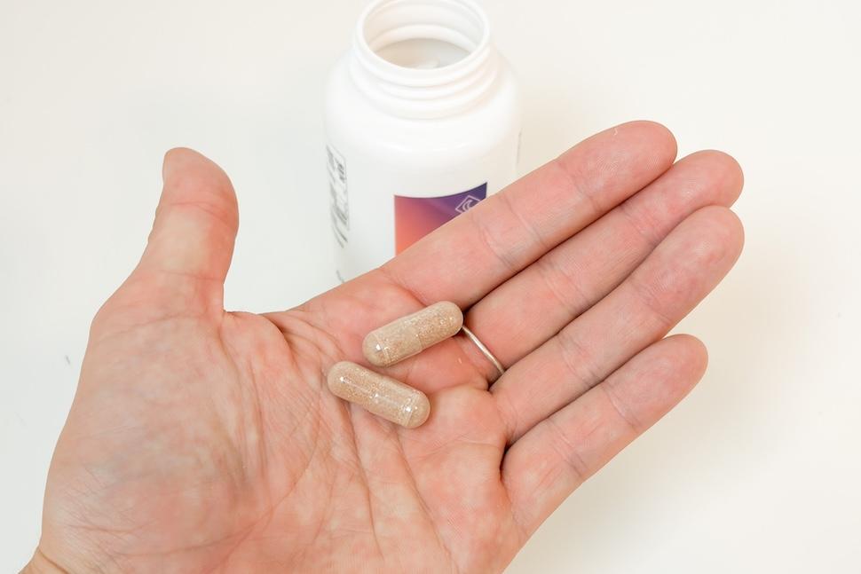 Closeup of a hand holding two Performance Lab Sleep capsules, which have a brownish color. You can see the open pill bottle on the countertop below.